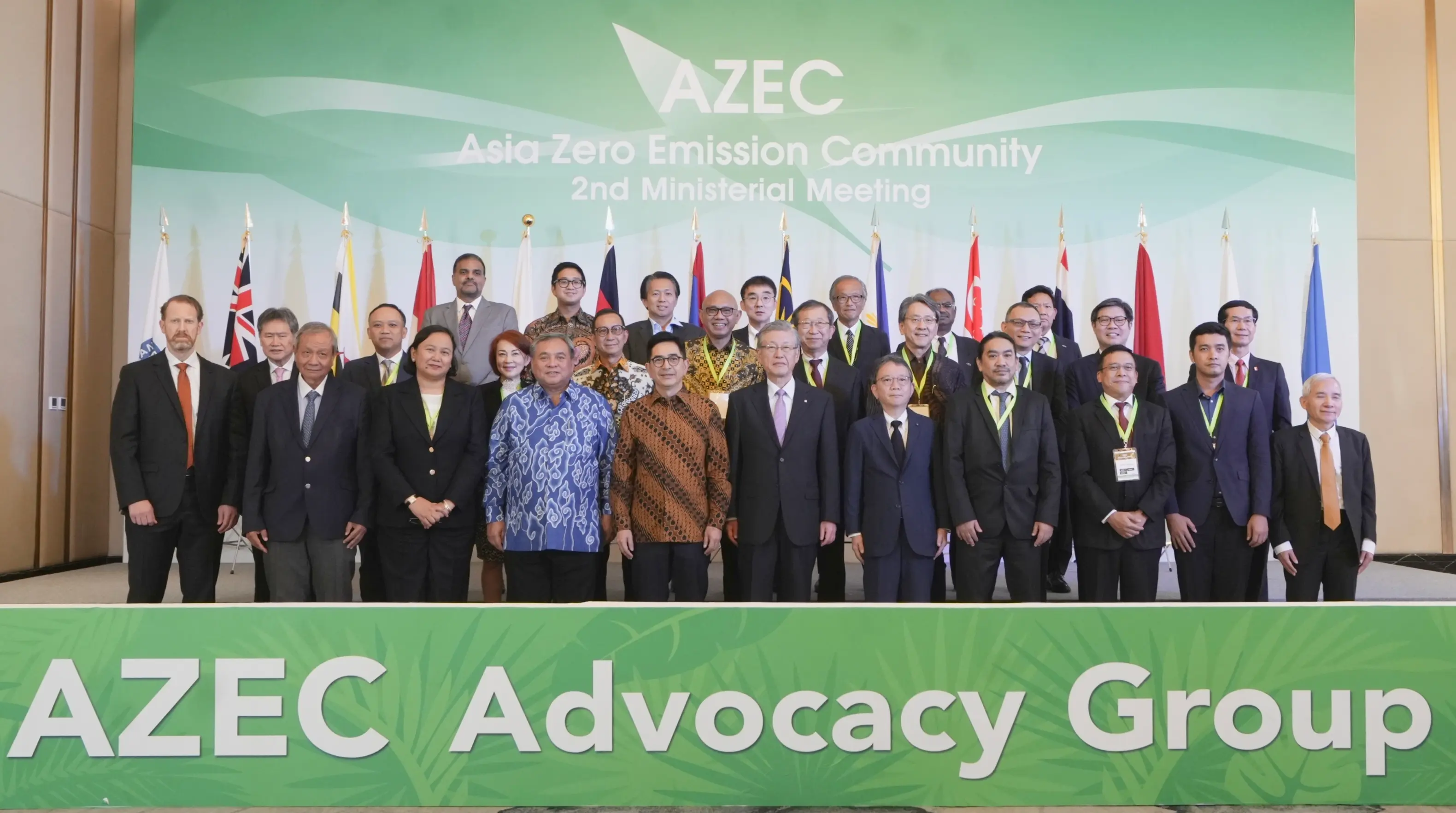 A group of diverse individuals stands together, dressed in formal attire, at the AZEC 2nd Ministerial Meeting. Various national flags are displayed behind them.