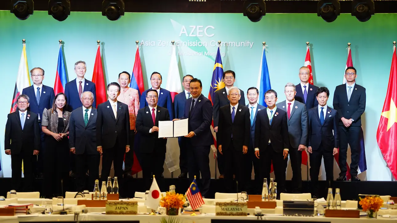 A group of diverse officials in suits stands in front of national flags at an international conference. Two individuals hold a signed document, symbolizing unity.