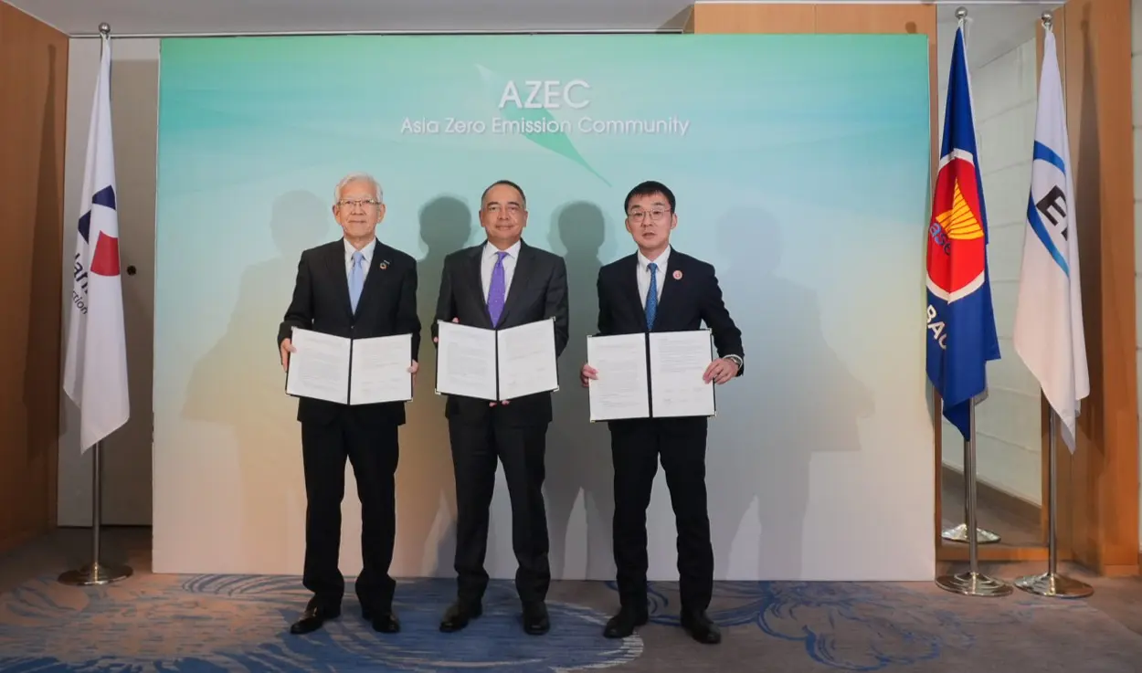 Three men in suits stand holding documents in front of a backdrop reading AZEC Asia Zero Emission Community, flanked by national flags, conveying a formal and cooperative tone.